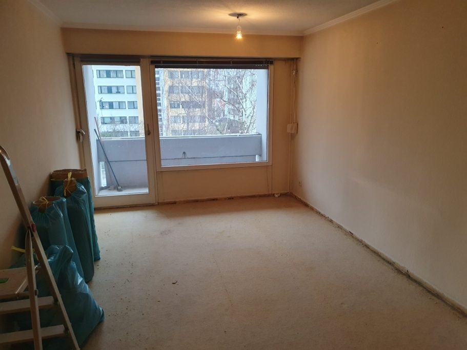 Leere Wohnraum mit Teppichboden, großen Fenstern und einem Balkon.