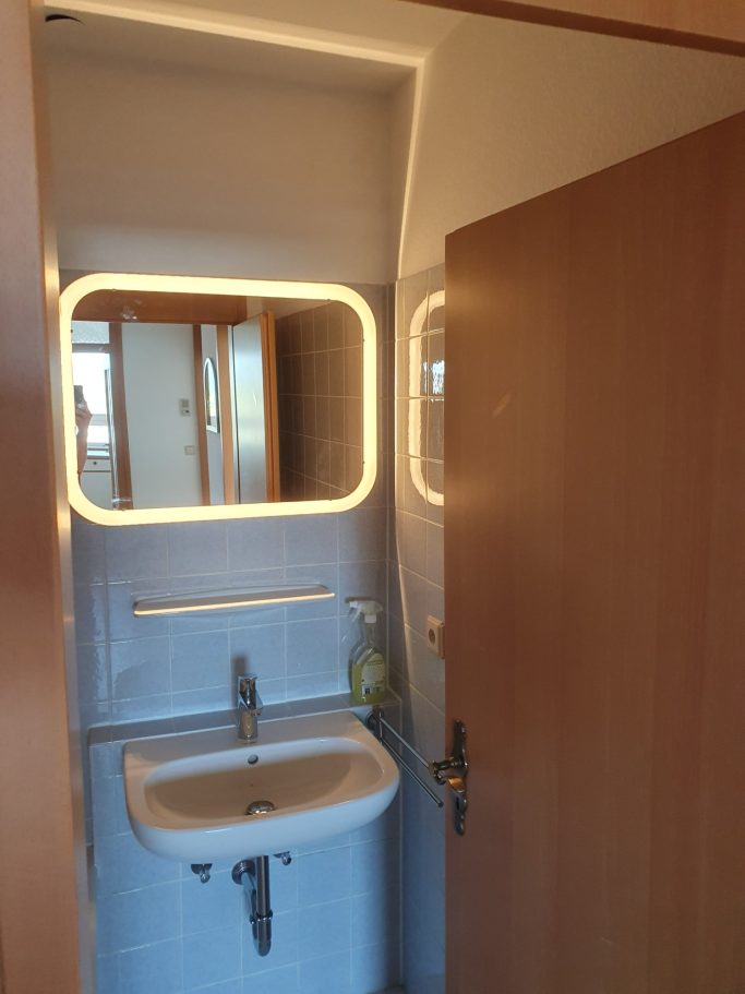 Moderne Toilette mit Waschbecken, beleuchtetem Spiegel und Holzfarbener Tür.