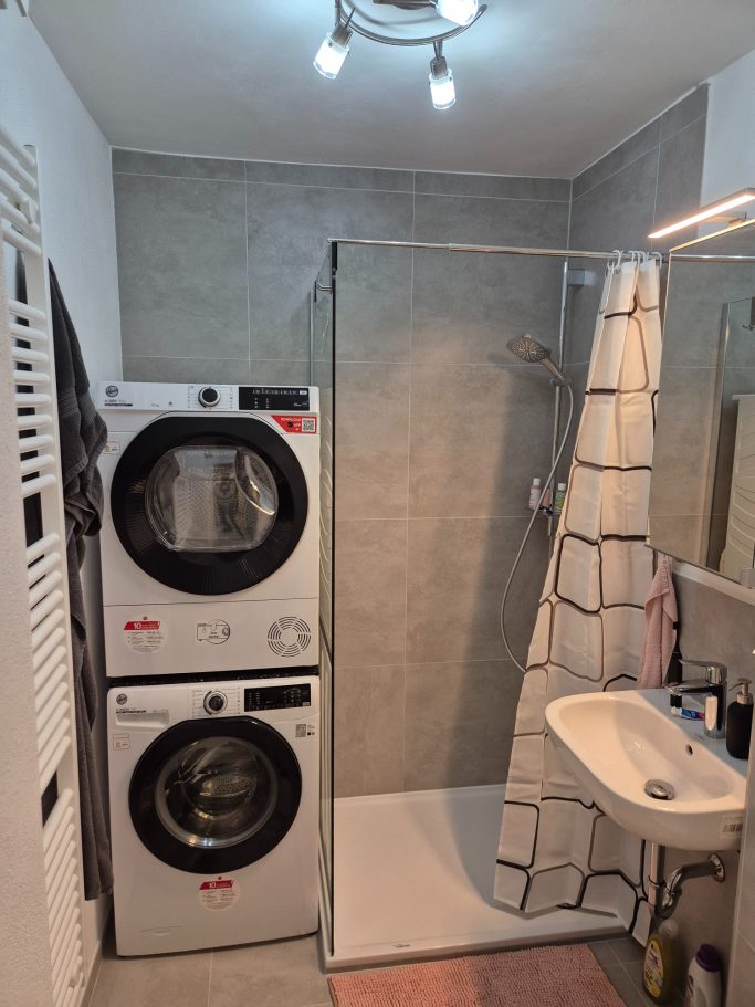 Kleines Badezimmer mit Waschmaschine, Trockner, Dusche und Waschbecken.