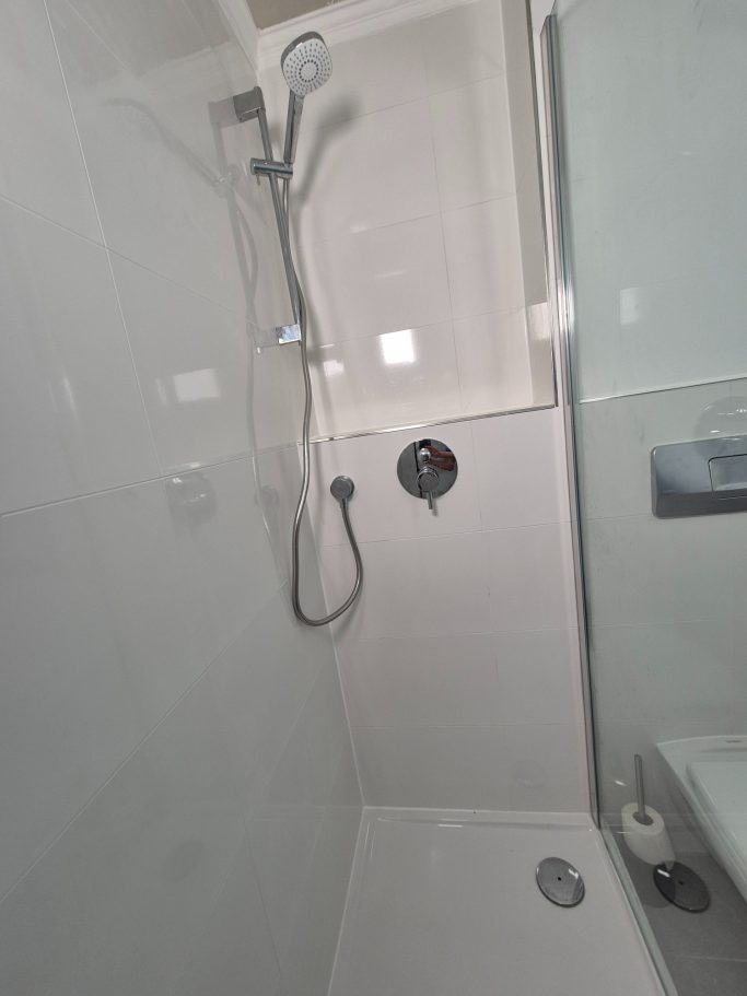 Badrenovierung Moderne Dusche mit Glaswand und verchromtem Duschkopf in hellen Fliesen.