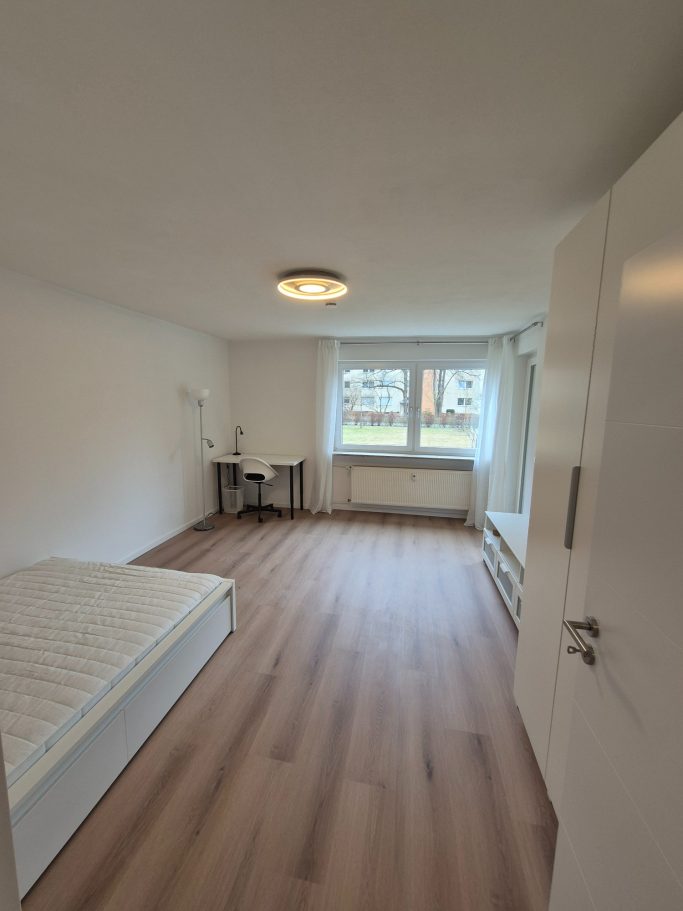 Helles Zimmer mit einem Bett, Schreibtisch und Fenster, holzähnlicher Boden.