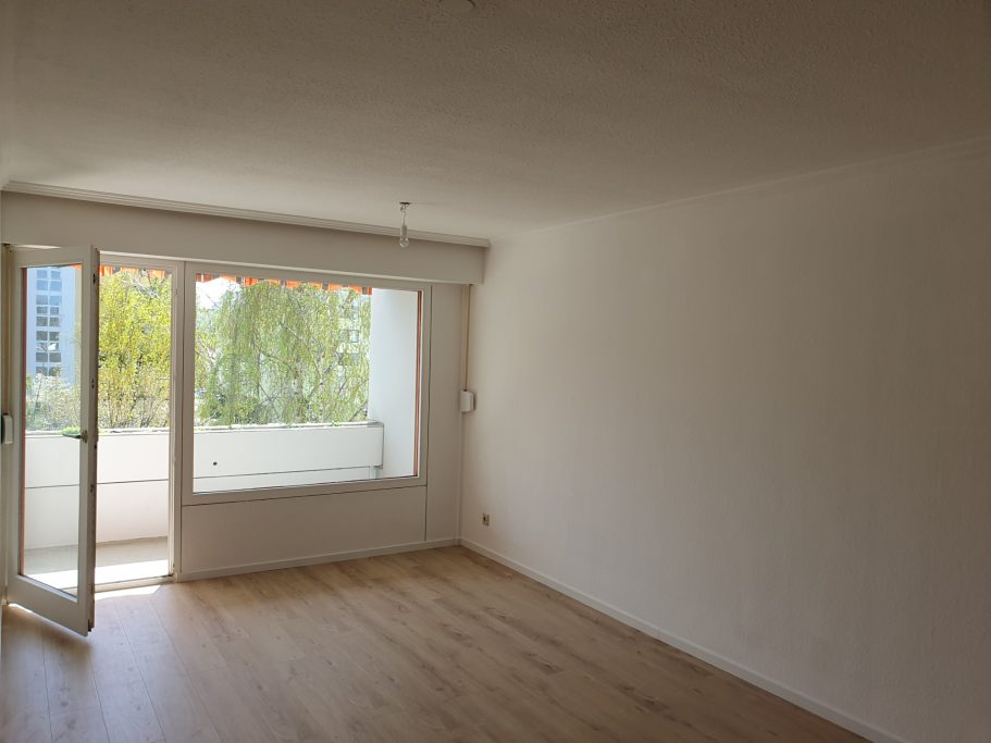 Helle, leere Wohnung mit großen Fenstern und Zugang zu einem Balkon.